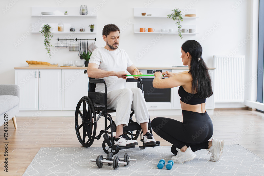 Handicapped mature man training with stretching bands. Concept of rehabilitation of disabled people. Young physiotherapist or wife in sportswear helping male patient in wheelchair exercise at home.