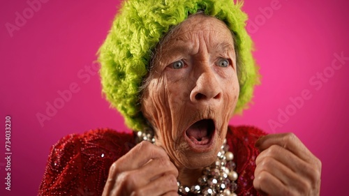 Surprised happy winner fisheye view of funny elderly woman with no teeth and green hat isolated on pink background. Caricature of peoples emotions.