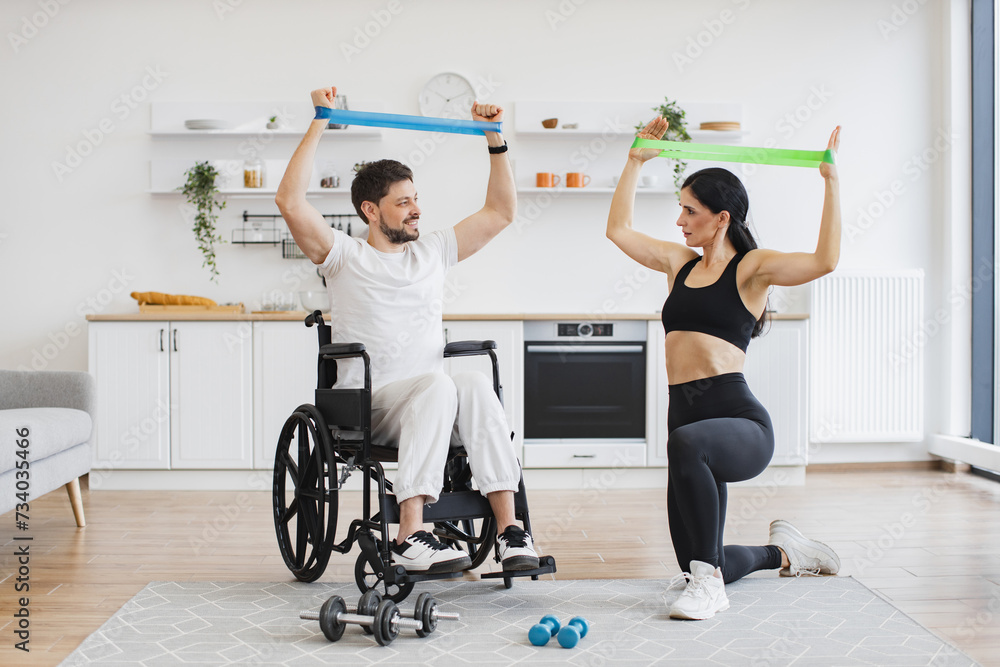 Fototapeta premium Wife in sportswear helping mature man in wheelchair lifting arms with stretching bands for muscle strength. Physiotherapy healthcare, medical trainer consulting disabled caucasian patient.
