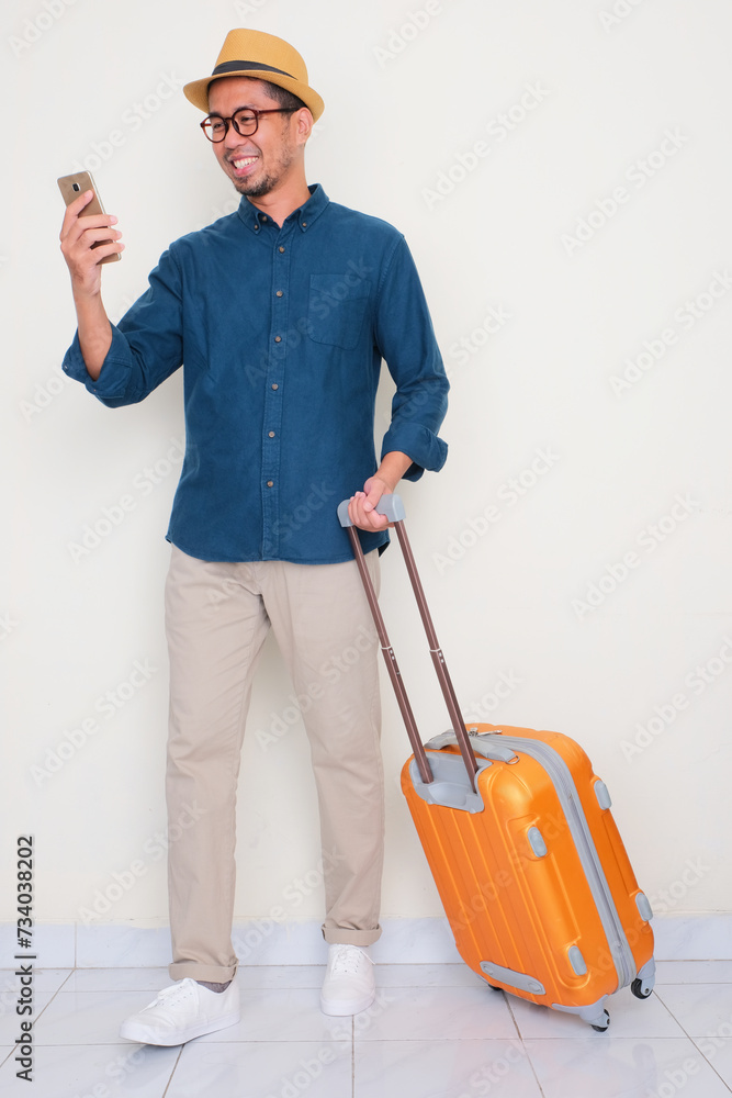 Full body portrait of a man pulling suitcase and looking to mobile phone with happy expression Full body portrait of a man pulling suitcase and looking to mobile phone with happy expression