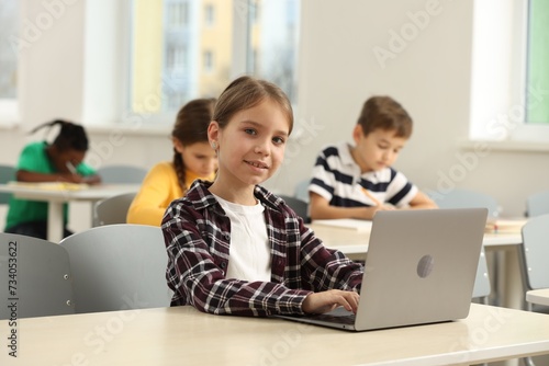 Smiling little girl with laptop studying in classroom at school