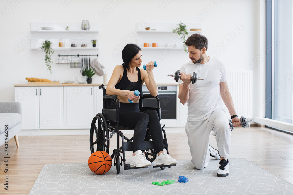 Rehab fitness for senior patients concept. Male physiotherapist or husband showing mature female patient in wheelchair how to exercise with dumbbells at home. - obrazy, fototapety, plakaty 