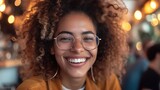 Close-up of a smiling young woman with curly hair and stylish glasses, embodying joy and positivity in a bustling cafe environment.