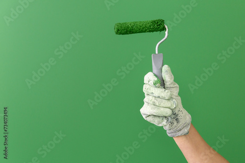 Close up horizontal image of hands in working gloves holding middle-sized roller with green paint in front of green wall.