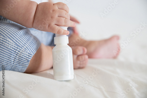 Hand of little child holding empty nasal spray bottle with space for text on background of legs sitting on white bed