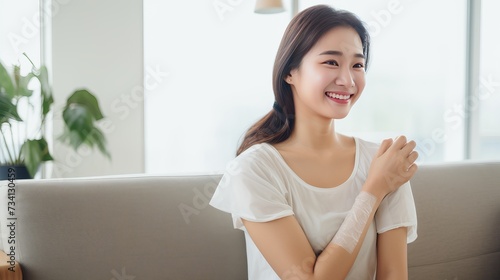 Happy Asian woman sitting on sofa showing arm with plaster after vaccination