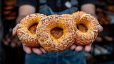 A pair of hands holding three large, sesame and poppy seed-covered pretzels in front of a bakery.