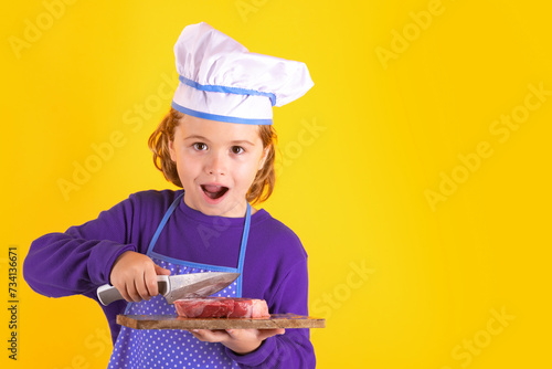 Kid cook hold cutting board with meat beef steak and knife. Child in cook uniform. Chef kid isolated on yellow background. Cute child to be a chef. Child dressed as a chef hat.