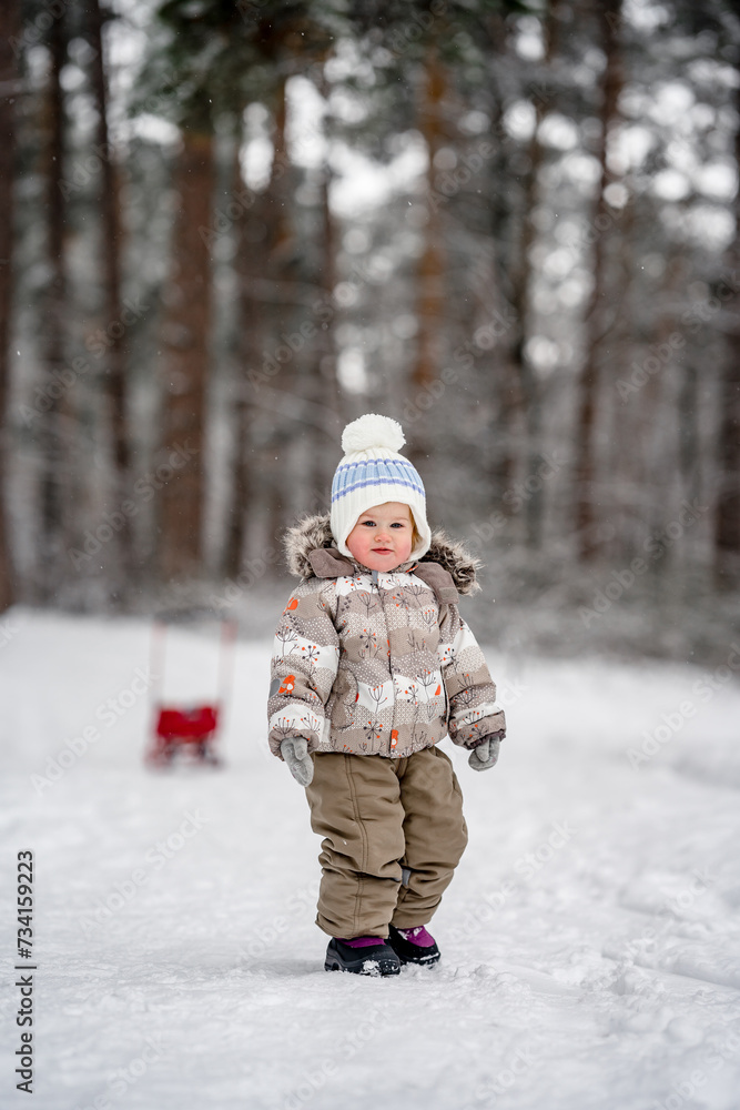 Little baby child in a warm jumpsuit walks in a snowy winter forest