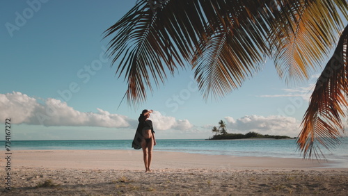 Woman walking on exotic tropical beach and looking away. Tourist girl in black bikini and shirt enjoy palm tree leaf, sea and wild island landscape. Outdoor lifestyle travel on summer holiday vacation