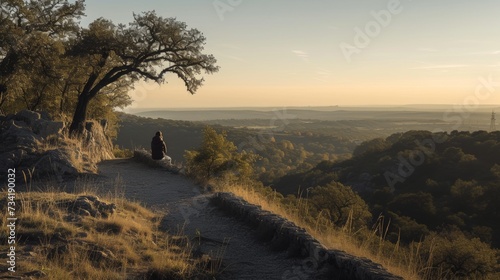 Individual sitting in solitude on a hill, overlooking a tranquil valley with a river snaking through it at dusk.