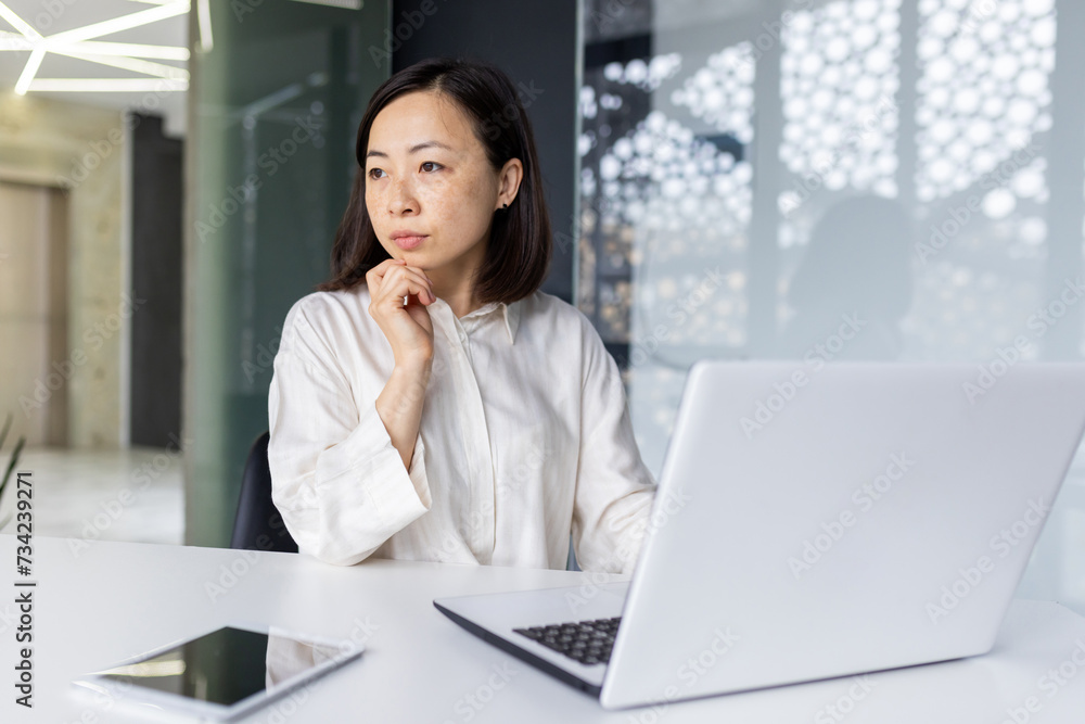 Photo of a successful and serious Asian business woman sitting in the office at a desk, working on a laptop and looking thoughtfully to the side. Photo of a successful and serious Asian business woman sitting in the office at a desk, working on a laptop and looking thoughtfully to the side.