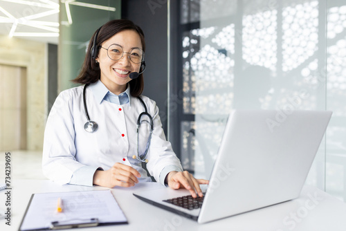 Portrait of a smiling young female Asian doctor sitting in the clinic at the work table in a headset and white coat in front of a laptop, working remotely, looking confidently into the camera.