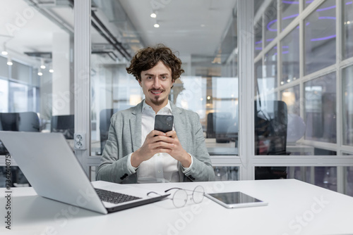 Portrait of a smiling and successful young businessman man in a suit sitting at a desk and using a mobile phone, looking confidently at the camera.