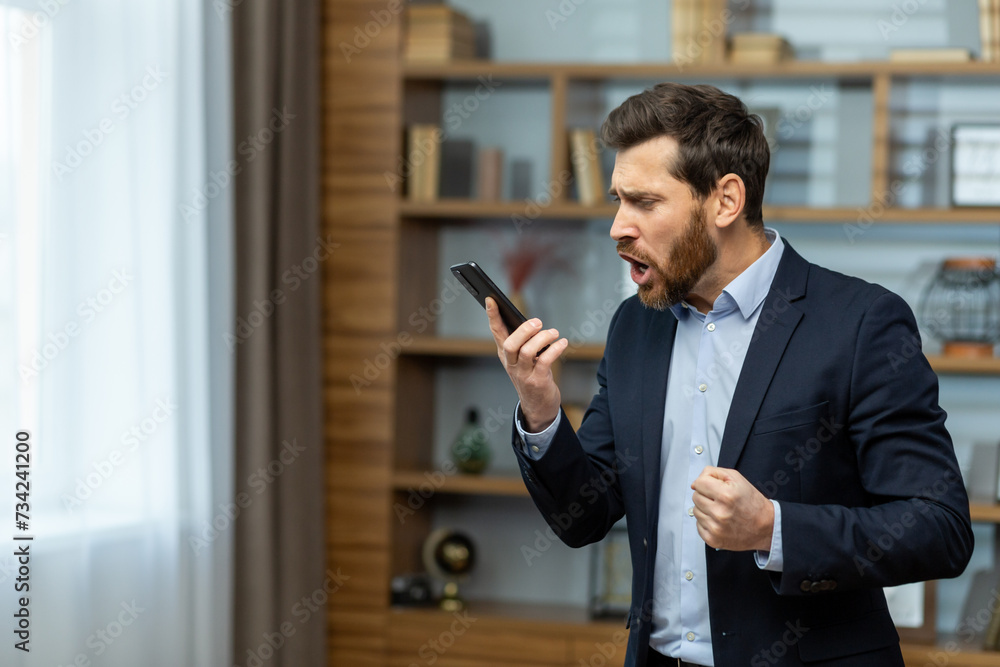 Angry young male businessman standing in the office in a suit and emotionally talking on the phone. Holds a mobile phone and shouts into the loudspeaker, solves questions and problems. Angry young male businessman standing in the office in a suit and emotionally talking on the phone. Holds a mobile phone and shouts into the loudspeaker, solves questions and problems.