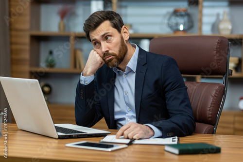 A young businessman man in a suit sits in the office at a desk in front of a laptop, is sad and upset, leaning his head and hand. He looks thoughtfully to the side.