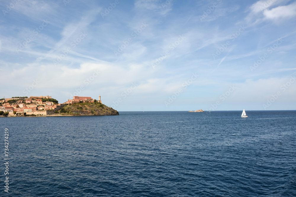 Seascape on the far shore with a lighthouse and ancient houses located on the hills. A yacht is sailing near the shore
