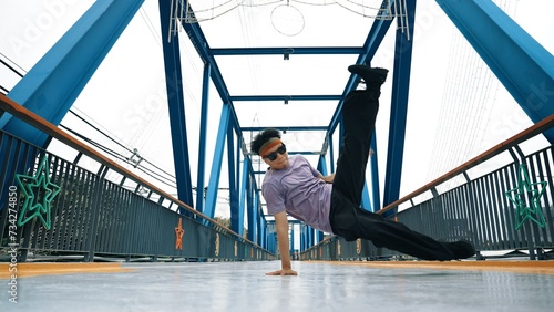 Hipster perform b-boy footstep and street dance at bridge with background. Professional break dancer wear stylish cloth and looking at camera while pose at camera. Outdoor sport 2024. Sprightly.