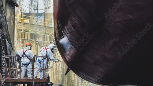 Shipyard Painters wearing personal protective clothing, gas masks and hazmat suits apply paint to keel of a large ship