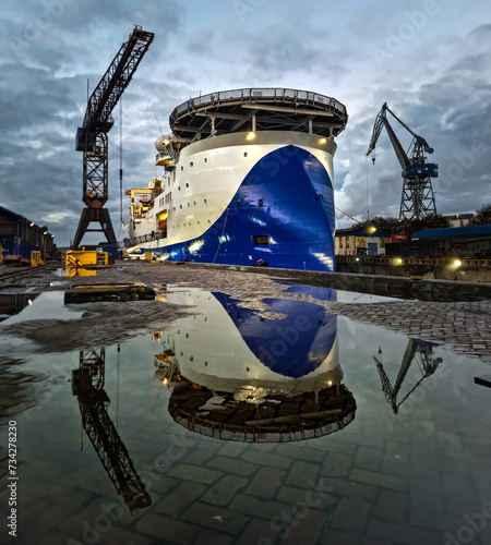 Large seismic offshore research ship in dry dock after painting hull surrounded with industrial cranes with water reflection
