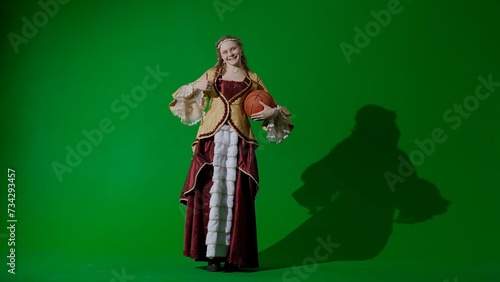 Woman in ancient outfit on the chroma key green screen background. Female in renaissance style dress holding basketball ball smiles shows thumb up.