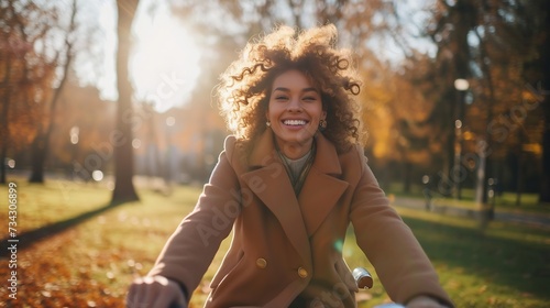 Generative AI : Smiling woman with curly hair in a coat rides a bicycle in a sunny park. Outdoor portrait. Beautiful woman enjoys nature.  © The Little Hut