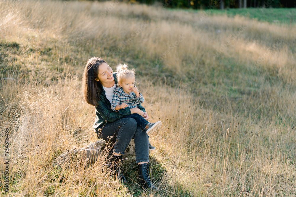 Smiling mother with a little girl on her knees sits on a stone in a sunny meadow