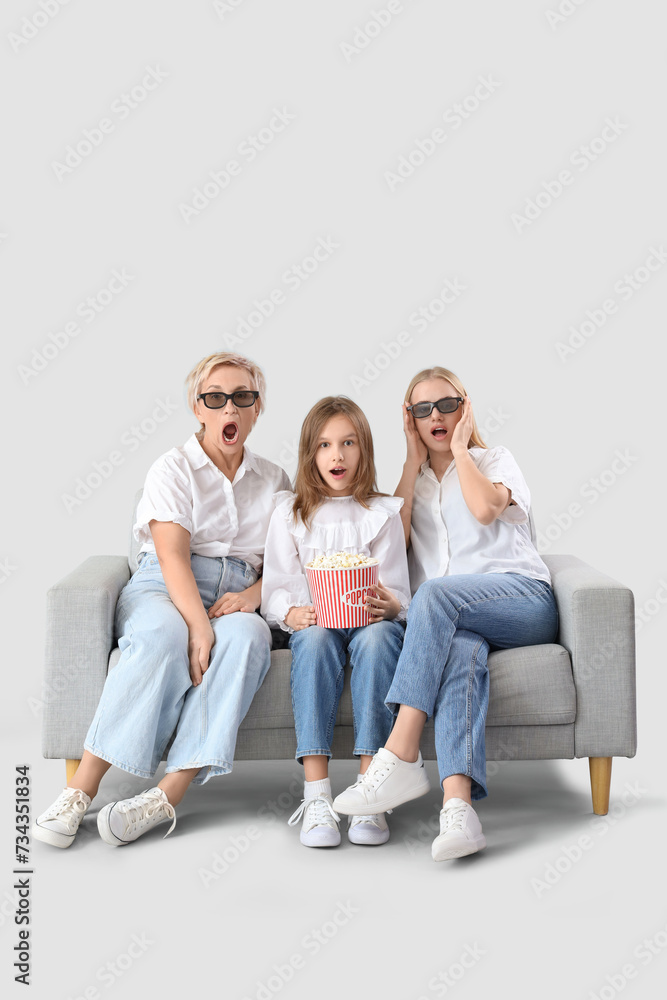Fototapeta premium Shocked little girl with her mom and grandmother watching movie on sofa against light background