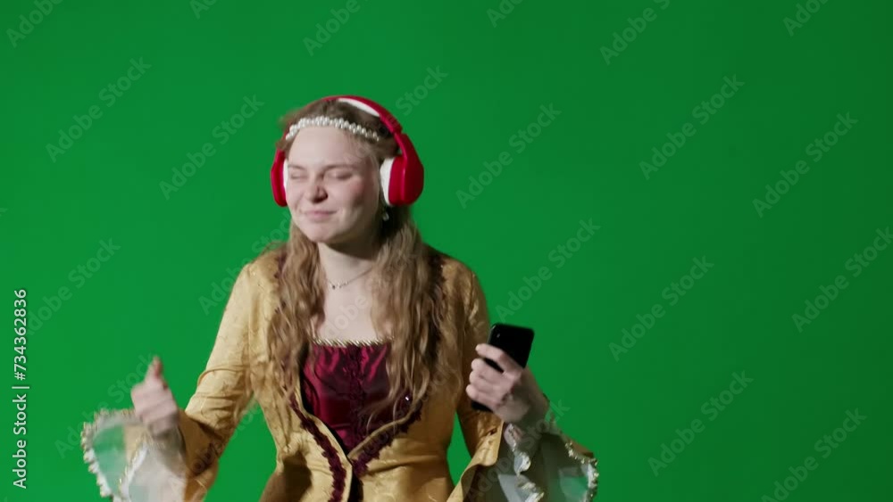 Woman in ancient outfit on chroma key green screen. Female in renaissance style dress in headphones listens music on smartphone and dancing. Woman in ancient outfit on chroma key green screen. Female in renaissance style dress in headphones listens music on smartphone and dancing.