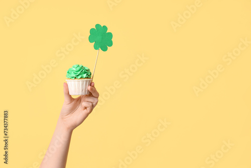 Female hand with tasty cupcake and paper clover for St. Patrick's Day on beige background