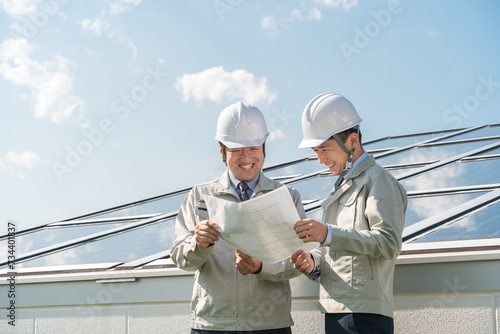 Foto 太陽光発電・ソーラーパネルの前に立つ電力会社・電気工事業者の作業着姿の技術者（クリーンエネルギー）