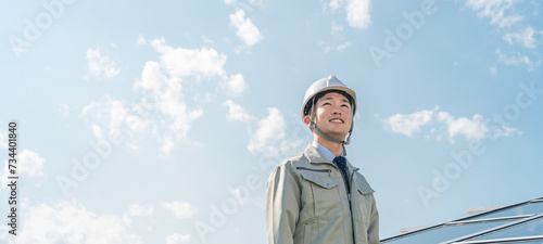 Foto 太陽光発電・ソーラーパネルの前に立つ電力会社・電気工事業者の作業着姿の技術者（クリーンエネルギー）