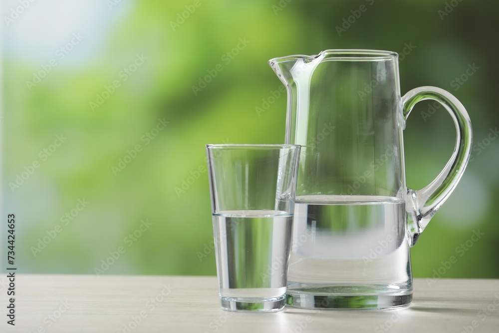 Jug and glass with clear water on white table against blurred green background, closeup. Space for text Jug and glass with clear water on white table against blurred green background, closeup. Space for text