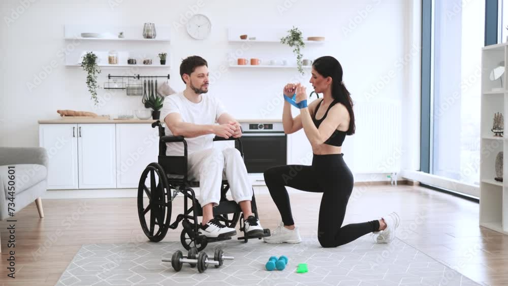Concept of rehabilitation of disabled people. Young physiotherapist or wife in sportswear helping male patient in wheelchair exercise at home. Handicapped mature man training with stretching bands . Concept of rehabilitation of disabled people. Young physiotherapist or wife in sportswear helping male patient in wheelchair exercise at home. Handicapped mature man training with stretching bands .