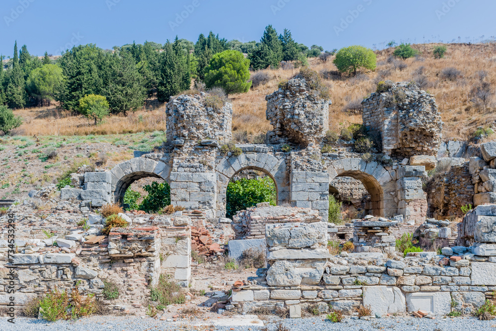 Stone ruins with iconic arches set in a dry grassy landscape under a bright blue sky in Ephesus, Turkiye.