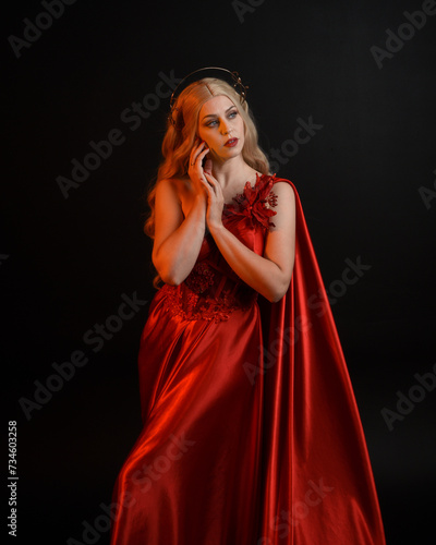 Close up portrait of beautiful blonde model wearing flowing red silk toga gown and crown, dressed as ancient mythological fantasy goddess. Graceful elegant pose isolated on dark studio background.