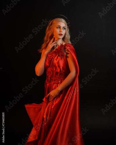 Close up portrait of beautiful blonde model wearing flowing red silk toga gown and crown, dressed as ancient mythological fantasy goddess. Graceful elegant pose isolated on dark studio background.