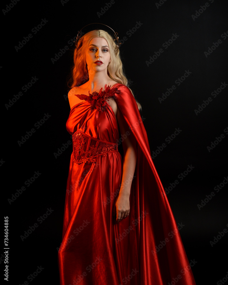 Naklejka premium Close up portrait of beautiful blonde model wearing flowing red silk toga gown and crown, dressed as ancient mythological fantasy goddess. Graceful elegant pose isolated on dark studio background.