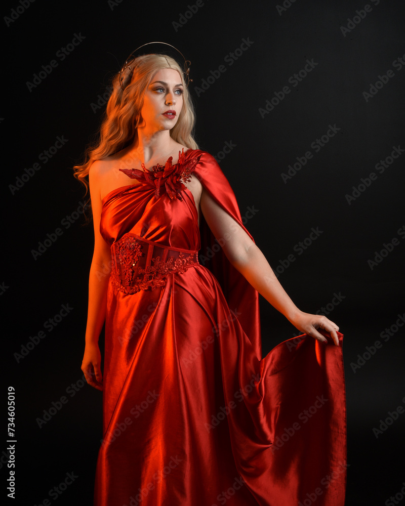Close up portrait of beautiful blonde model wearing flowing red silk toga gown and crown, dressed as ancient mythological fantasy goddess. Graceful elegant pose isolated on dark studio background.