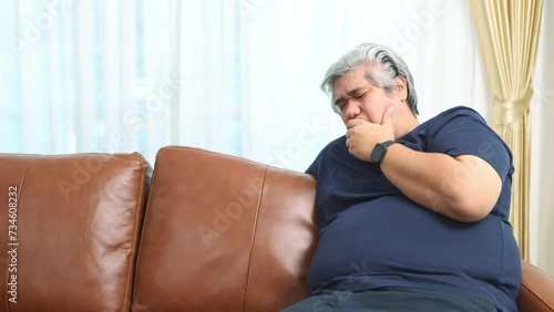 Obese asian man sit on the sofa has health problems suffering from respiratory and lung diseases chronic cough severe chronic bronchitis lung infection symptoms choking on food or saliva. photo
