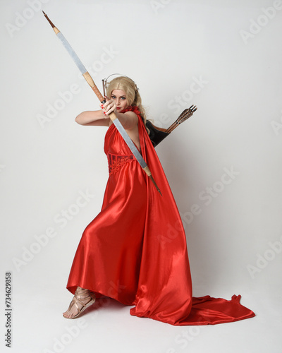 Full length portrait of blonde model dressed as mythological fantasy goddess in flowing red silk toga gown, crown. Graceful elegant pose holding archery weapons, isolated on studio background