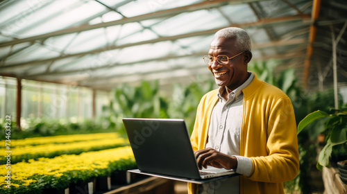 A dark-skinned African American man with a gadget, laptop, tablet is planting and caring for plants, seedlings, vegetables in a greenhouse.