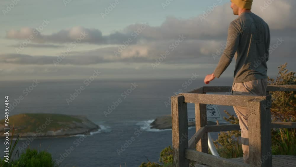 Man walks onto a wooden lookout platform, looking into the rising sun in the Torndirrup national park, medium framing, walks out of shot. Man walks onto a wooden lookout platform, looking into the rising sun in the Torndirrup national park, medium framing, walks out of shot.