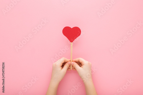 Little child hands holding red paper heart shape on wooden stick on light pink table background. Pastel color. Love, health and hope concept. Closeup. Top down view. Point of view shot.