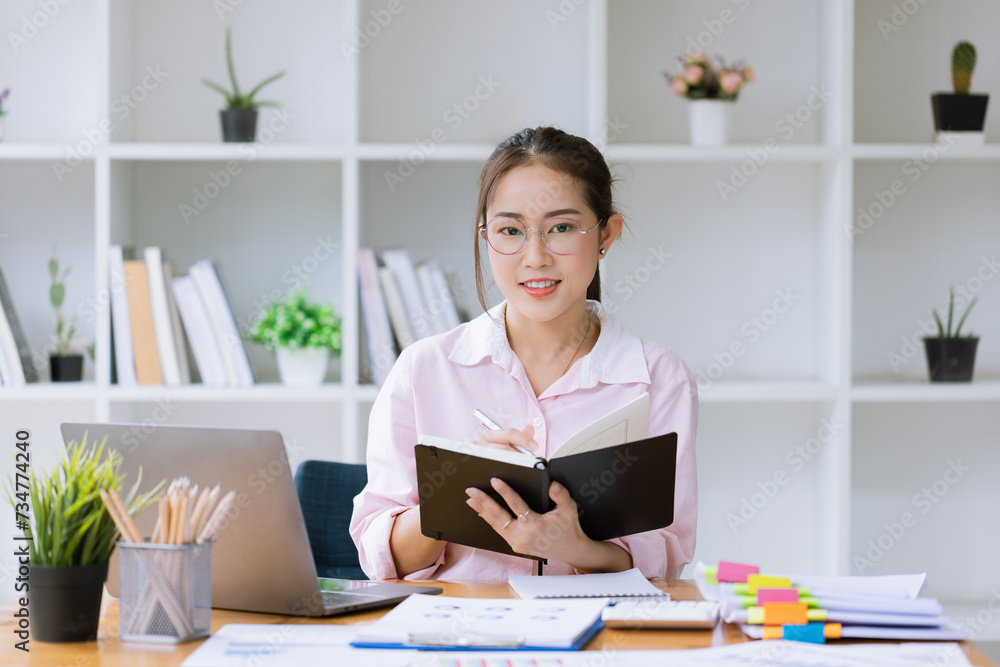 Young asian woman wear glasses reading a book with laptop sitting at desk in the study room. Young asian woman wear glasses reading a book with laptop sitting at desk in the study room.