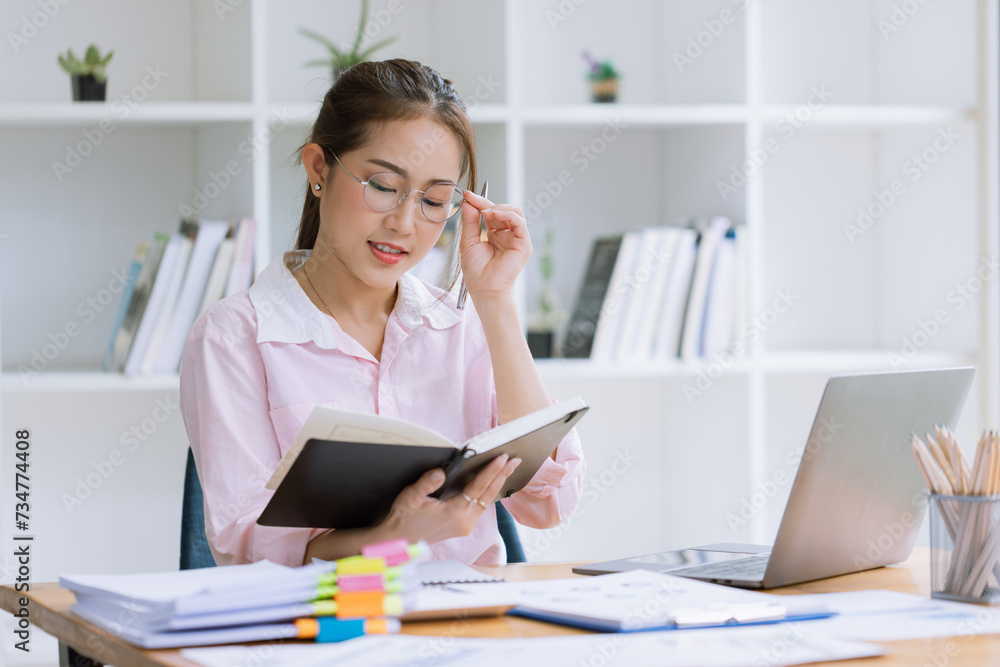 Young asian woman wear glasses reading a book with laptop sitting at desk in the study room. Young asian woman wear glasses reading a book with laptop sitting at desk in the study room.