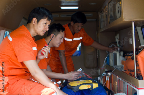 Two male nurse in an orange uniform measure vital signs of an accident victim on a bed in an ambulance. The other man use walkie talkie to coordinate the nearest hospital.