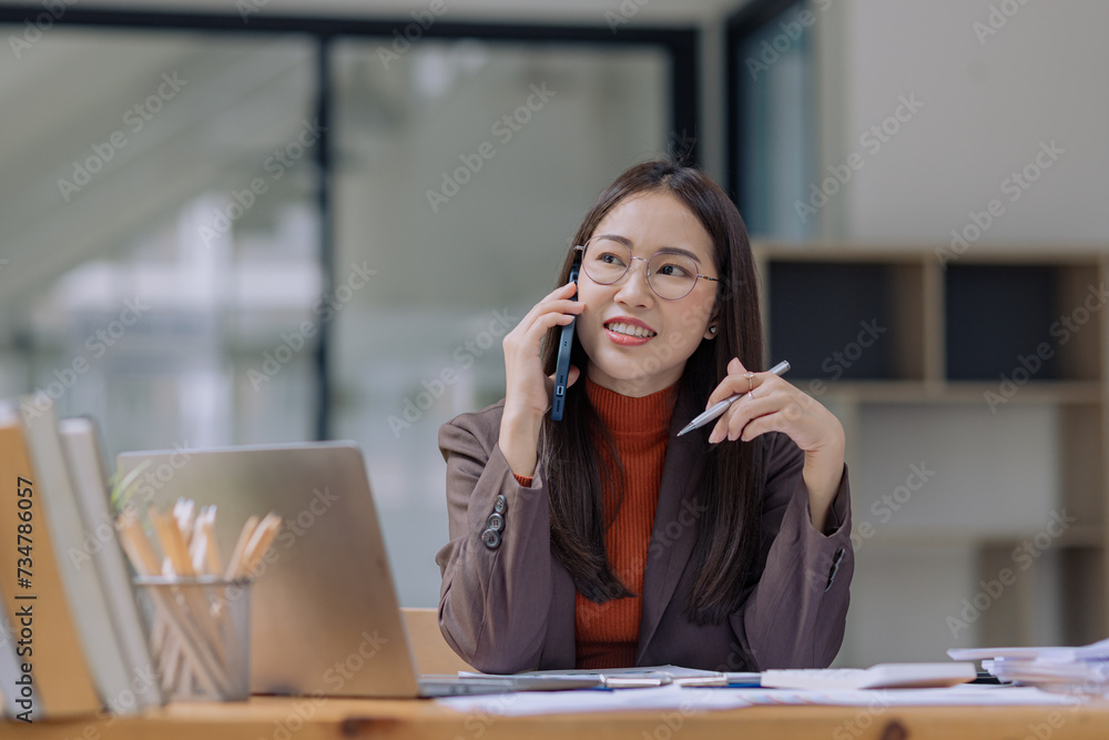 Happy asian young businesswoman using digital smartphone sitting in office working space, Asian female employee using laptop talking on the phone at workplace. Happy asian young businesswoman using digital smartphone sitting in office working space, Asian female employee using laptop talking on the phone at workplace.