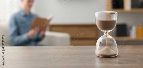 Hourglass with flowing sand on desk. Man reading book in room, selective focus