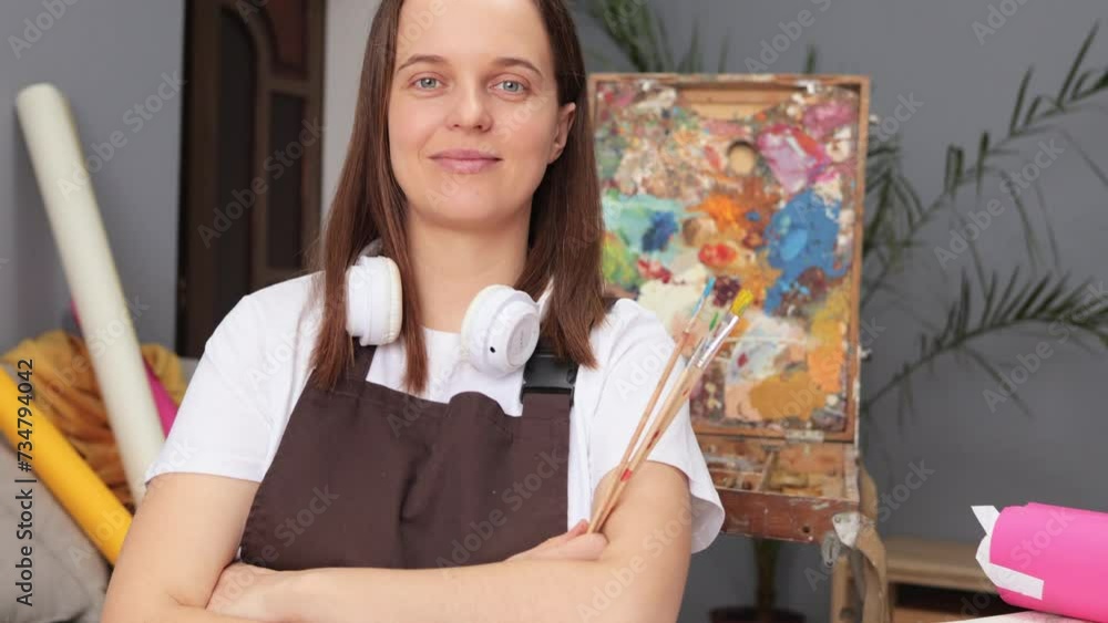 Smiling cheerful Caucasian brown haired woman wearing apron sitting at workplace in drawing studio enjoying her hobby creating works of art. Imaginative art exploration. Smiling cheerful Caucasian brown haired woman wearing apron sitting at workplace in drawing studio enjoying her hobby creating works of art. Imaginative art exploration.